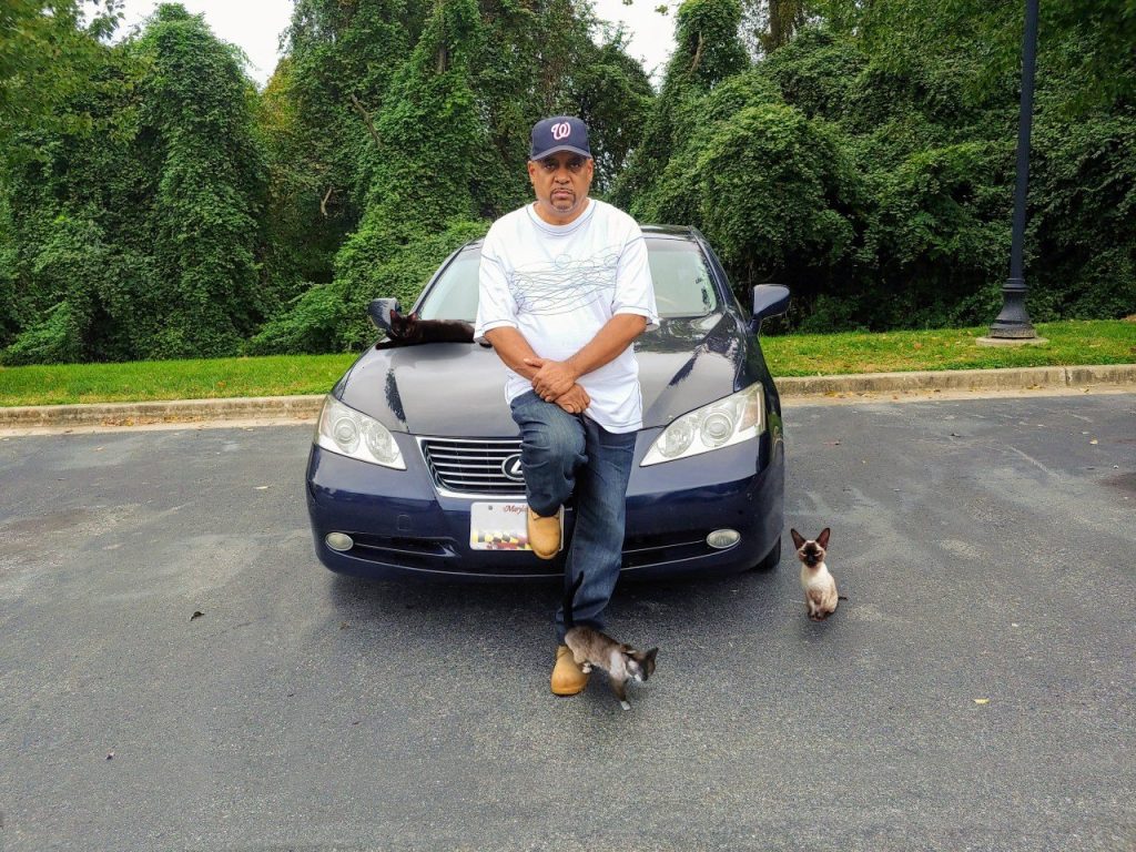 A man in a cap leans against a parked car. Two small dogs are on the ground nearby. Trees are in the background.